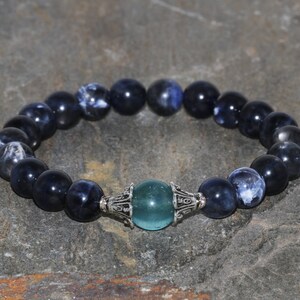 Puede incluir: Un brazalete de piedras azules y blancas con un cierre plateado y una cuenta de piedra verde turquesa en el centro.