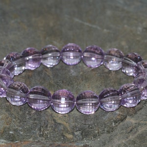 Op de afbeelding: Een armband gemaakt van facetgeslepen, lichtpaarse kralen. De ronde kralen hebben een gesneden patroon dat licht reflecteert. De armband is elastisch en rekt uit om de pols te passen. De achtergrond is een grijze stenen ondergrond.
