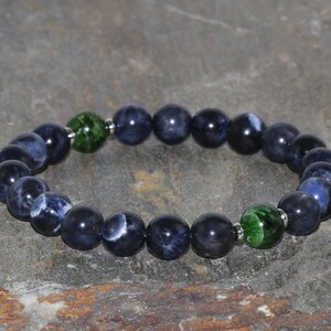 Puede incluir: Un brazalete de piedras azules y blancas con detalles verdes. El brazalete está hecho de cuentas redondas y tiene un cierre plateado.