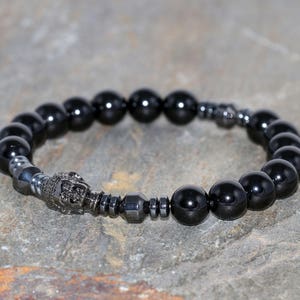 Puede incluir: Pulsera de cuentas negras con un dije de Buda plateado y cuentas de hematita.