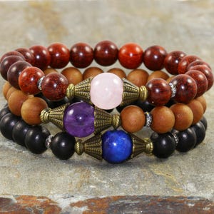 Puede incluir: Tres pulseras de cuentas apiladas. La pulsera superior presenta cuentas de madera de color rojo oscuro. La pulsera del medio tiene cuentas de madera de color marrón claro y una piedra preciosa rosa. La pulsera inferior tiene cuentas negras y una gema púrpura.