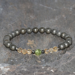 Puede incluir: Un brazalete de piedra de pirita gris con una cuenta de aventurina verde y cuentas doradas.