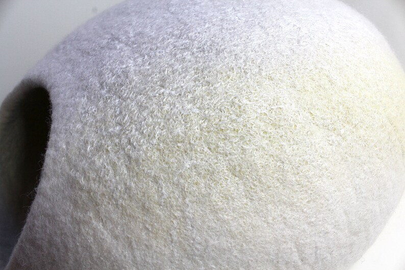 May include: A white felt cat bed shaped like a cave. The bed has a soft, fuzzy texture and a light yellow hue.