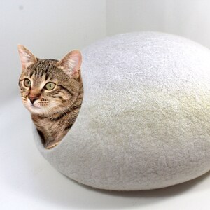 May include: A brown tabby cat with green eyes peeks out of a white felt cat bed shaped like an egg.