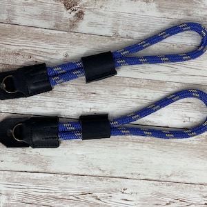 Może przedstawiać: Dwa niebiesko-białe paski do aparatu z liną, z czarnymi skórzanymi końcami i metalowymi pierścieniami. Paski leżą na jasnej powierzchni z usłojeniem drewna.
