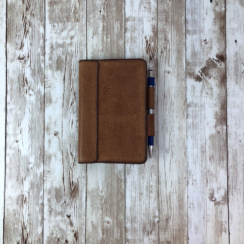 K&ouml;nnte beinhalten: Ein braunes, ledergebundenes Notizbuch mit einem Stift, der an der Abdeckung befestigt ist. Das Notizbuch liegt auf einer wei&szlig; get&uuml;nchten Holzoberfl&auml;che.