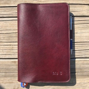 K&ouml;nnte beinhalten: Ein burgunderfarbener Leder-Notizbuchumschlag mit einem Stift, der in die Seitentasche gesteckt ist. Der Umschlag tr&auml;gt die Initialen "Mis S" auf der Vorderseite.