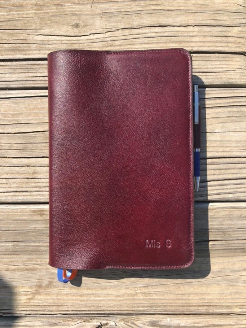 K&ouml;nnte beinhalten: Ein burgunderfarbener Leder-Notizbuchumschlag mit einem Stift, der in die Seitentasche gesteckt ist. Der Umschlag tr&auml;gt die Initialen "Mis S" auf der Vorderseite.