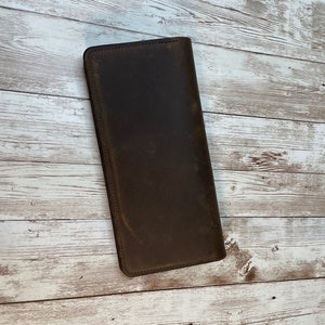 May include: A brown leather wallet with a distressed finish. The wallet is closed and lying flat on a white wooden surface.
