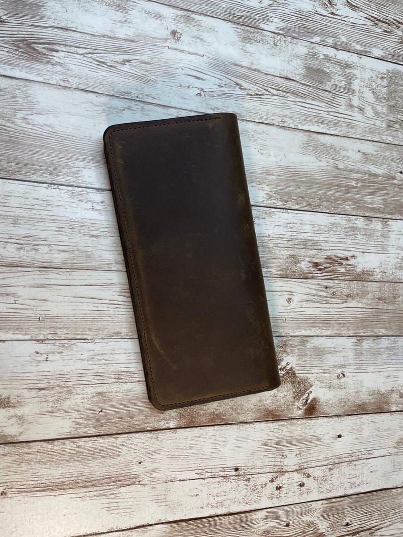 May include: A brown leather wallet with a distressed finish. The wallet is closed and lying flat on a white wooden surface.