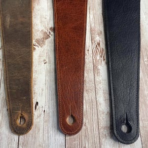 May include: Three leather guitar straps in varying shades of brown and black. The straps are made of textured leather and have a hole at the bottom for attaching to a guitar. The straps are displayed on a light-colored wooden surface.