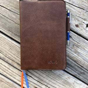 K&ouml;nnte beinhalten: Ein braunes Lederjournal mit einem Stift, der in die Seitentasche gesteckt ist. Das Journal hat ein Lesezeichen aus orangefarbenem und blauem Band und die Initialen "KMJ" sind auf dem Einband gepr&auml;gt.