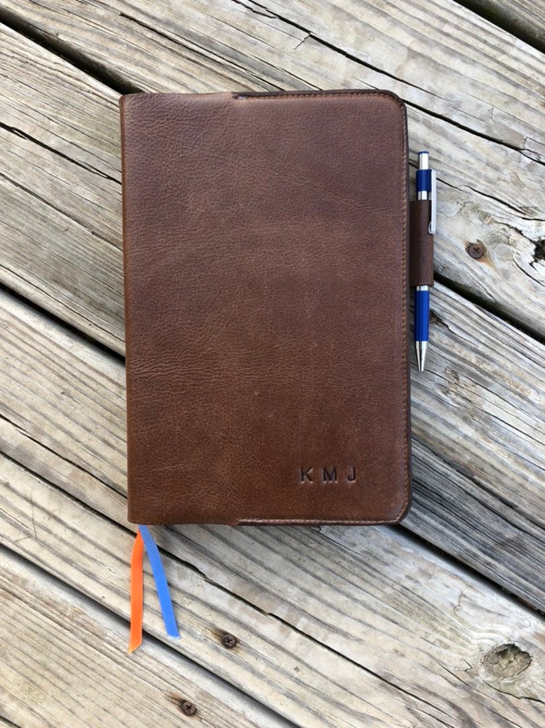 K&ouml;nnte beinhalten: Ein braunes Lederjournal mit einem Stift, der in die Seitentasche gesteckt ist. Das Journal hat ein Lesezeichen aus orangefarbenem und blauem Band und die Initialen "KMJ" sind auf dem Einband gepr&auml;gt.