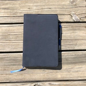 K&ouml;nnte beinhalten: Ein schwarzes Notizbuch mit einem blauen Band-Lesezeichen und einem silbernen Stift. Das Notizbuch liegt auf einer Holzoberfl&auml;che.
