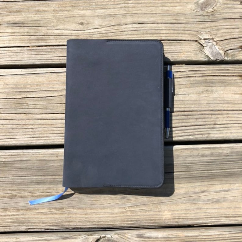 K&ouml;nnte beinhalten: Ein schwarzes Notizbuch mit einem blauen Band-Lesezeichen und einem silbernen Stift. Das Notizbuch liegt auf einer Holzoberfl&auml;che.