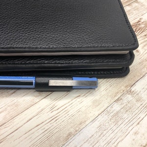 May include: A black leather-bound notebook with a blue pen on top of it. The notebook is closed and has a white edge.