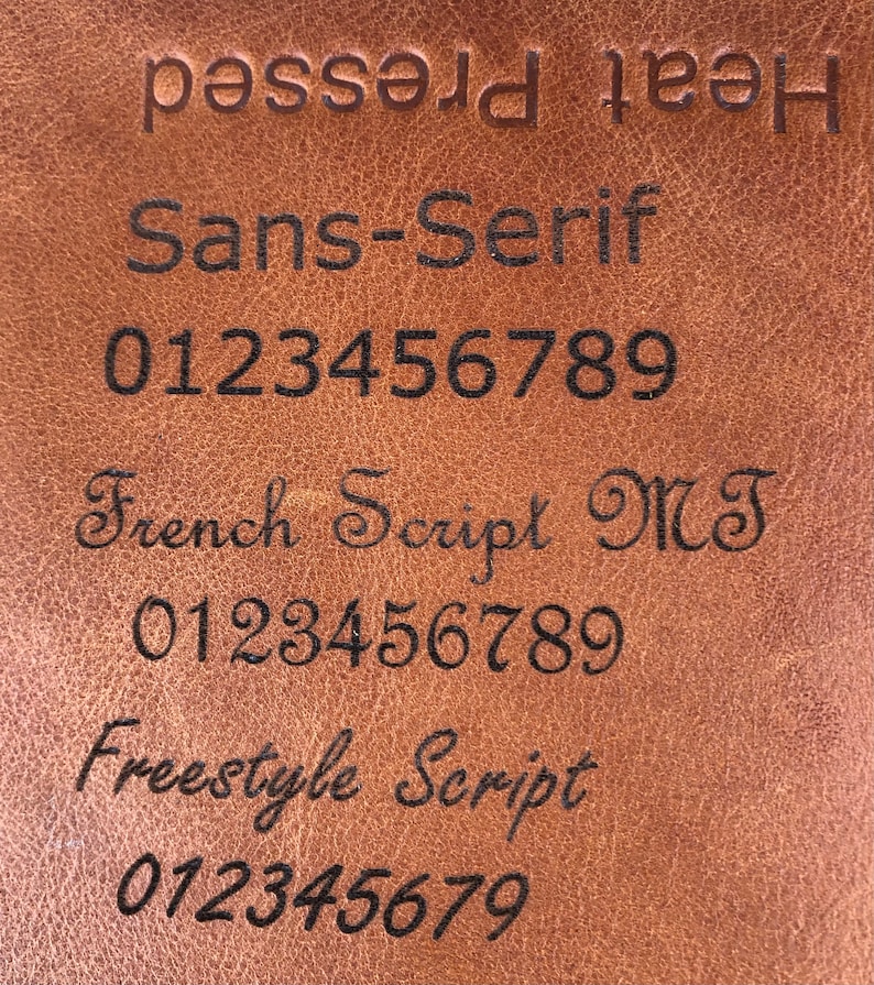 May include: A close-up of a brown leather surface with the text "Heat pressed" at the top. Below the text are three different font styles: Sans-Serif, French Script MT, and Freestyle Script. Each font style is accompanied by the numbers 0123456789.