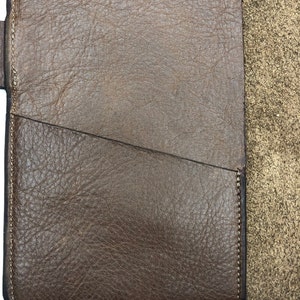 K&ouml;nnte beinhalten: Nahaufnahme einer braunen Leder-Notizbuchh&uuml;lle mit einem gen&auml;hten Rand und einer Tasche auf der Vorderseite. Die H&uuml;lle ist aus strukturiertem Leder mit einem dezenten Glanz.