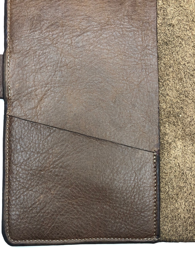 K&ouml;nnte beinhalten: Nahaufnahme einer braunen Leder-Notizbuchh&uuml;lle mit einem gen&auml;hten Rand und einer Tasche auf der Vorderseite. Die H&uuml;lle ist aus strukturiertem Leder mit einem dezenten Glanz.