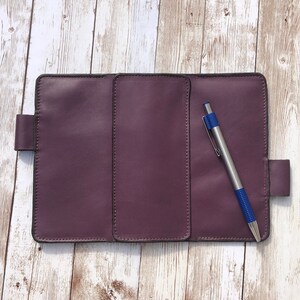 K&ouml;nnte beinhalten: Ein purpurfarbenes, ledergebundenes Tagebuch mit drei Abschnitten und einem blauen Stift. Das Tagebuch hat einen Klappverschluss und ein gen&auml;htes Design.