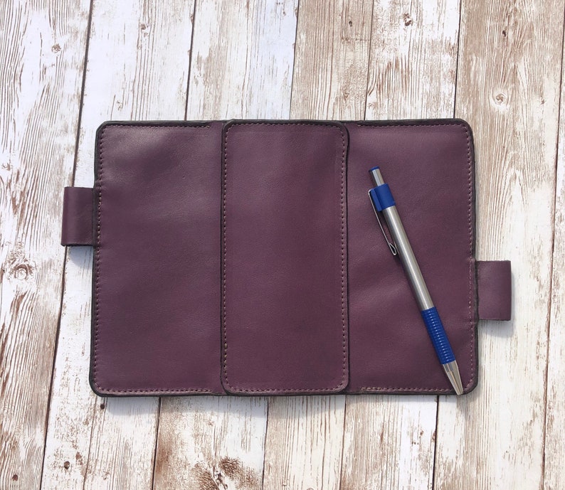 K&ouml;nnte beinhalten: Ein purpurfarbenes, ledergebundenes Tagebuch mit drei Abschnitten und einem blauen Stift. Das Tagebuch hat einen Klappverschluss und ein gen&auml;htes Design.
