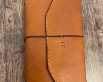 May include: A tan leather journal cover with a pointed flap closure secured by a thin black elastic band. The front of the cover features the embossed letters T J F. The leather has a smooth, matte finish with visible stitching along the edges.
