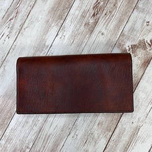 May include: A brown leather wallet with a smooth, textured surface. The wallet is closed and lying flat on a white wooden surface.