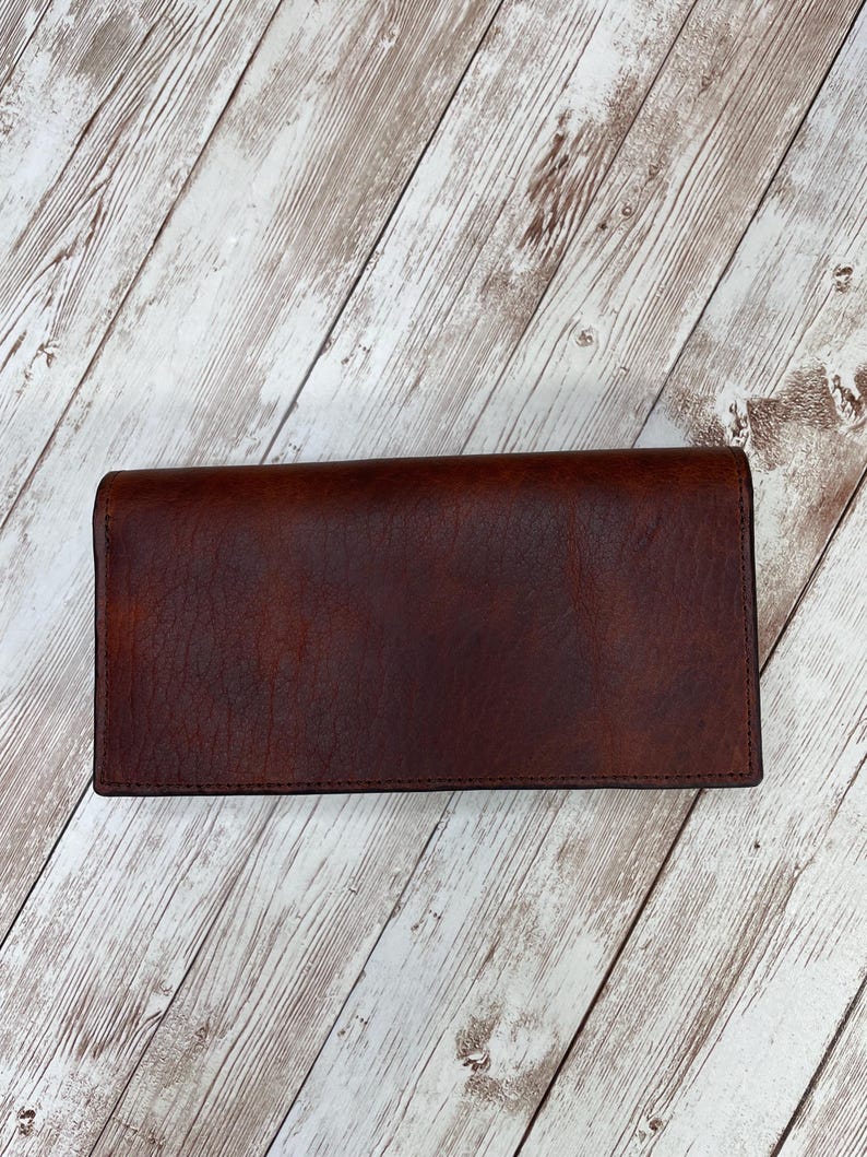 May include: A brown leather wallet with a smooth, textured surface. The wallet is closed and lying flat on a white wooden surface.
