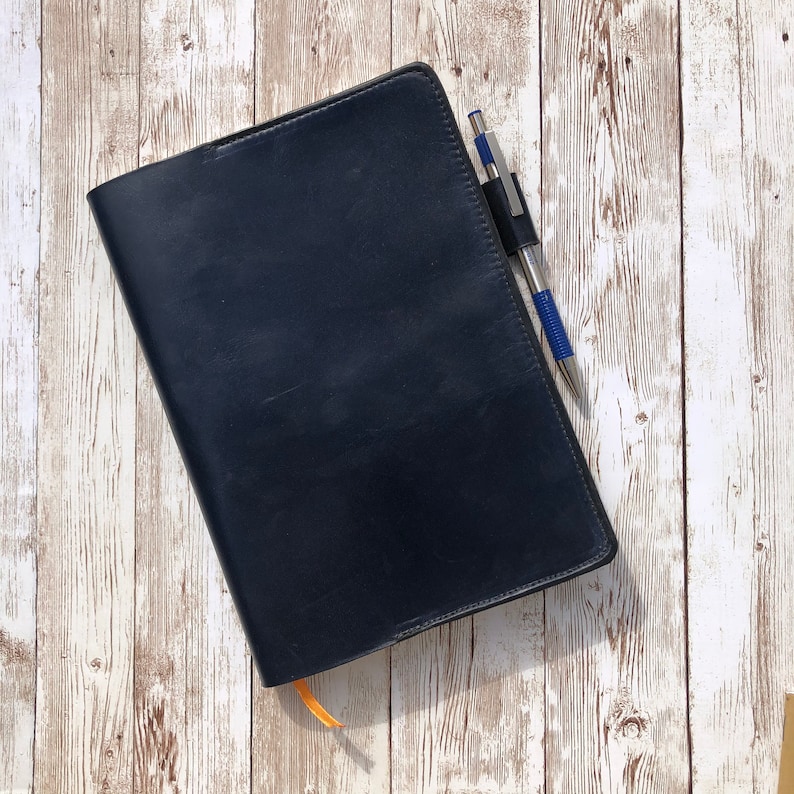 K&ouml;nnte beinhalten: Ein schwarzes, ledergebundenes Notizbuch mit einem silbernen Stift, der in die Umschlag gesteckt ist. Das Notizbuch liegt auf einer wei&szlig;en Holzoberfl&auml;che.