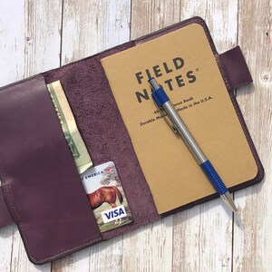 K&ouml;nnte beinhalten: Ein Notizbuchcover aus lila Leder mit einem Fach f&uuml;r Bargeld und Karten. Das Cover ist ge&ouml;ffnet und zeigt ein Field Notes Notizbuch mit einem silbernen Stift mit blauer Spitze.