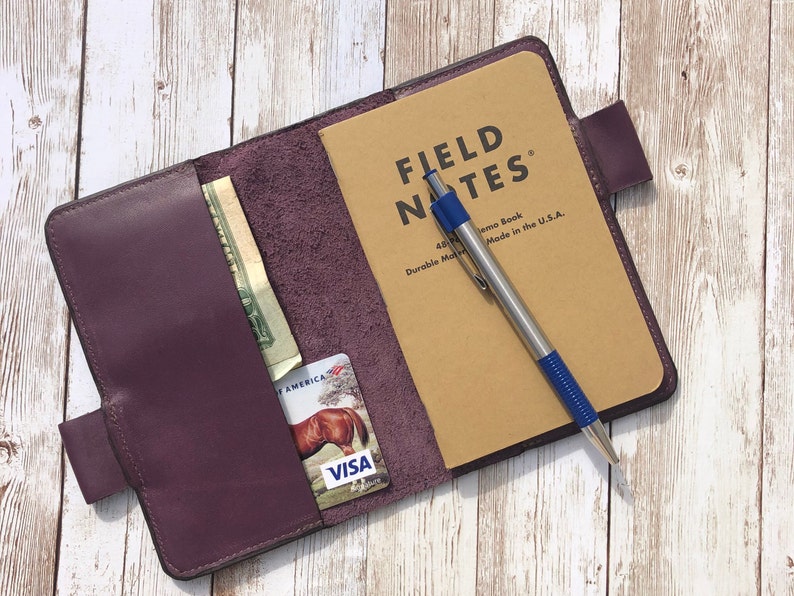 K&ouml;nnte beinhalten: Ein Notizbuchcover aus lila Leder mit einem Fach f&uuml;r Bargeld und Karten. Das Cover ist ge&ouml;ffnet und zeigt ein Field Notes Notizbuch mit einem silbernen Stift mit blauer Spitze.
