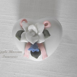May include: White porcelain heart-shaped trinket box with a white flower and pink ribbon decoration.
