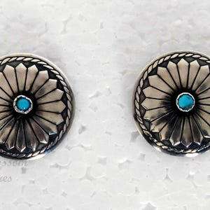 May include: Pair of silver-toned stud earrings. Each earring features a central turquoise-colored stone surrounded by a detailed, sunburst-like design. The earrings have a decorative border and are set against a white background.