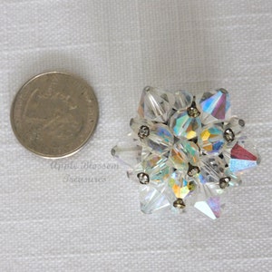 Puede incluir: Un grupo de cristales de vidrio transparente iridiscente, engastados en una base de metal plateado. Los cristales están dispuestos en forma de estrella. La pieza mide aproximadamente el tamaño de una moneda de 20 céntimos de euro.