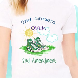 May include: White t-shirt with a graphic design of green sneakers with flowers, a sun, and clouds. The text "2nd Graders Over 2nd Amendment" is printed on the shirt.