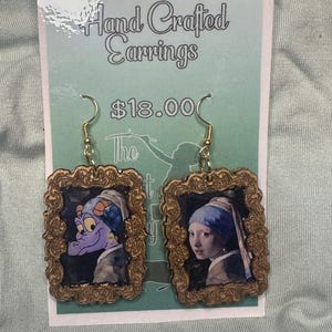 Peut inclure: Une paire de boucles d'oreilles artisanales avec un cadre doré orné d'un dragon violet et d'une fille portant un foulard bleu, tous deux dans un style de peinture. Les boucles d'oreilles coûtent 18,00 €.