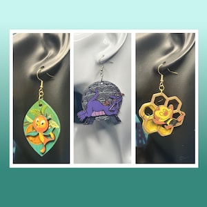 May include: Three pairs of novelty earrings. One pair features an orange character on a green leaf. Another pair showcases a purple dragon in front of a grey sphere. The third pair displays a yellow honeycomb with a cartoon character.