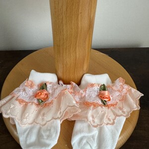 Puede incluir: Un par de calcetines blancos para bebé con ribete de encaje de color melocotón y un acento de flor de melocotón.