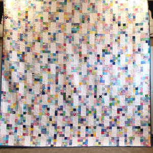 Puede incluir: Un edredón grande, hecho a mano, con un diseño de retazos. El edredón presenta un fondo blanco con una cuadrícula de cuadrados coloridos en varios tonos de azul, rosa, amarillo y verde. El edredón está bordeado con un fino ribete azul.