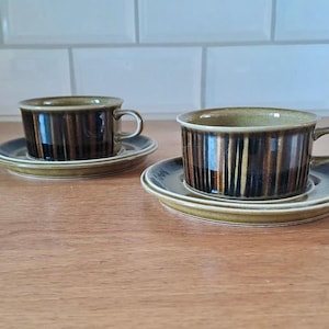 Peut inclure: Deux tasses à café et soucoupes assorties. Les tasses et les soucoupes sont marron avec des rayures verticales. Les soucoupes ont un bord vert clair. Les tasses ont une anse.