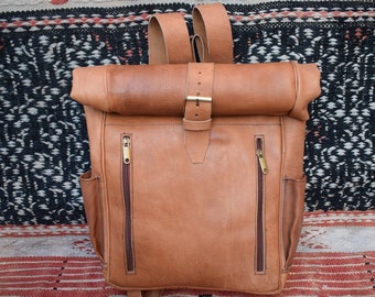 May include: A brown leather backpack with a roll-top closure and a buckle. The backpack has two zippered pockets on the front and two adjustable straps.