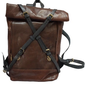 May include: A brown leather backpack with a roll-top and black straps. The backpack has adjustable straps with gold-coloured buckles. The design includes a front pocket and a top handle for carrying.