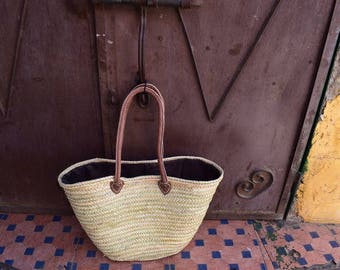 Puede incluir: Un bolso tote de paja tejida con asas y ribetes de cuero marrón. El bolso tiene un interior oscuro y está colocado contra una puerta marrón desgastada y un suelo de baldosas estampadas.