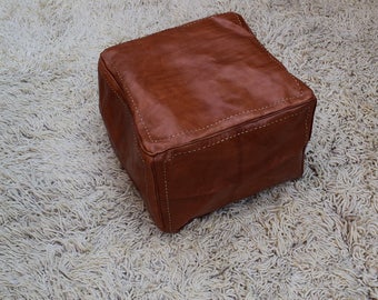 May include: A brown leather ottoman with white stitching. The ottoman is sitting on a white shag rug.