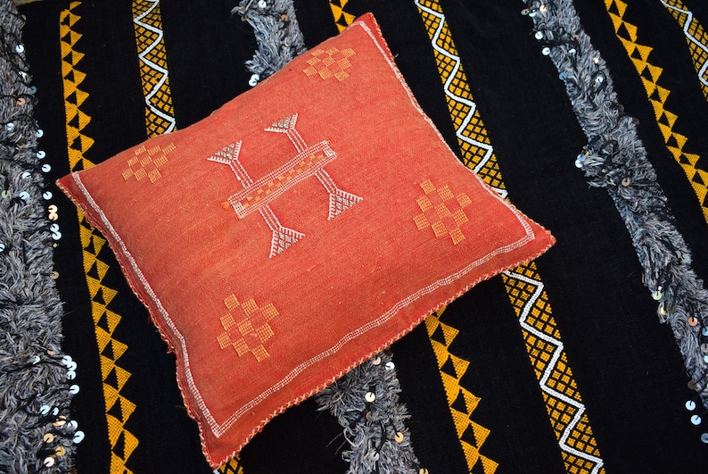 K&ouml;nnte beinhalten: Ein rotes Kissen mit wei&szlig;en N&auml;hten und einem geometrischen Muster. Das Kissen liegt auf einem schwarzen Stoff mit goldenen Mustern.