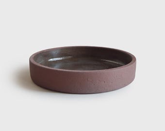 May include: A round, terracotta-colored ceramic dish with a dark brown interior. The dish has a slightly textured exterior.