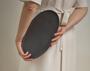 May include: A dark gray, oval-shaped serving platter. The platter is held in front of a person wearing a light-colored, linen-like dress. The platter has a smooth surface and a slightly raised edge.