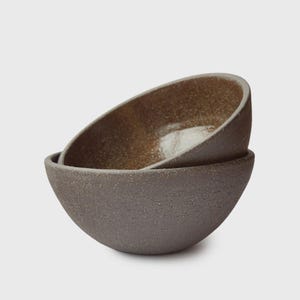 Puede incluir: Dos cuencos de cerámica, uno dentro del otro, con un exterior gris oscuro texturizado y un interior moteado de color bronce. Los cuencos tienen una forma redondeada y asimétrica, lo que sugiere un diseño artesanal.
