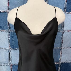 May include: A black slip dress with a cowl neckline and spaghetti straps. The dress is made of a shiny, silky material and hangs on a mannequin. The background is a patchwork of blue denim squares.