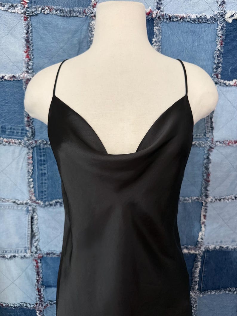 May include: A black slip dress with a cowl neckline and spaghetti straps. The dress is made of a shiny, silky material and hangs on a mannequin. The background is a patchwork of blue denim squares.